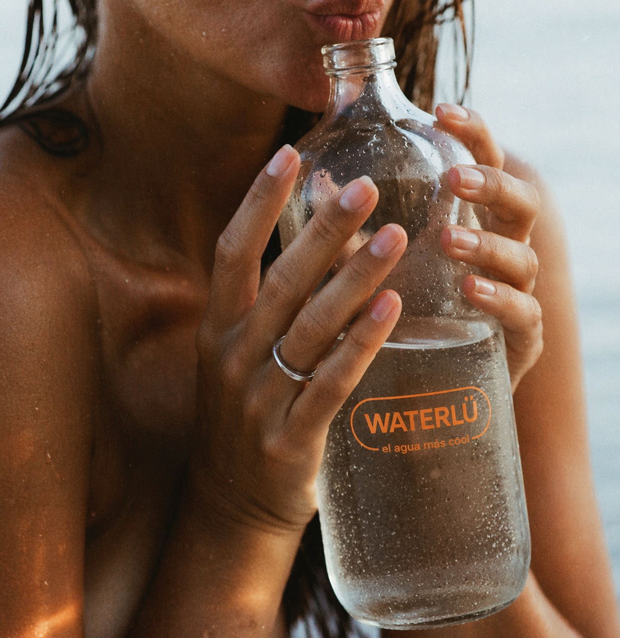 Manos sosteniendo botella WATERLÜ en la playa